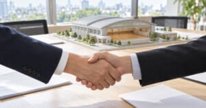 会議室で握手する2人の手元と体育館の模型