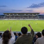 ナイター照明のサッカー場と歓声を上げる観客の後ろ姿