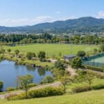 池のある公園の中の球技場とテニスコート、奥に低い山